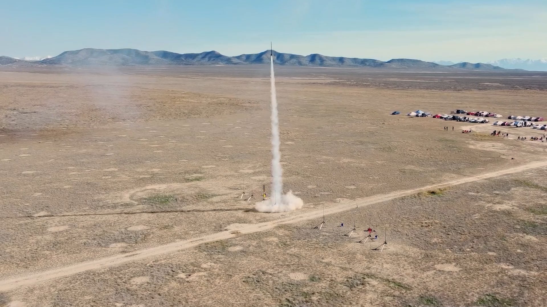 Aerial drone view of a rocket launching from a desert range, trailing a white smoke plume, with launch rails, parked cars and spectators nearby