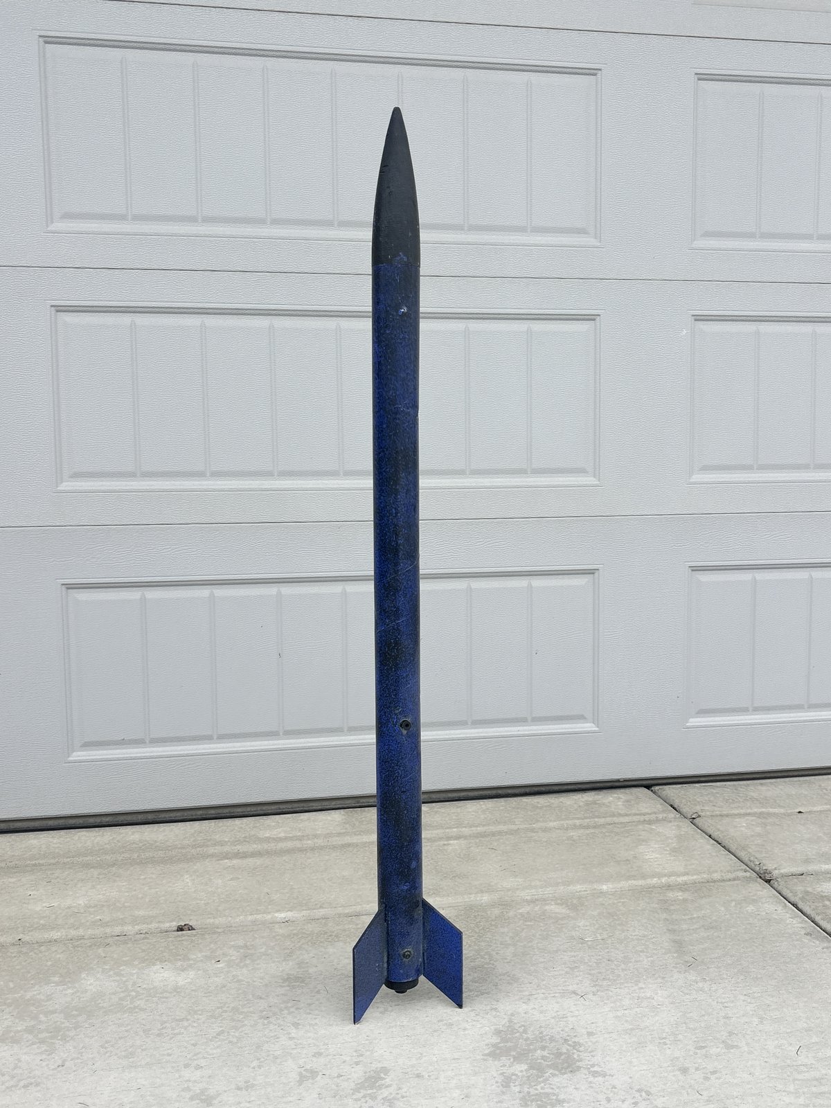 The finished rocket, tall and slender with a blue body, black nose cone and three blue fins, standing upright in front of a garage door