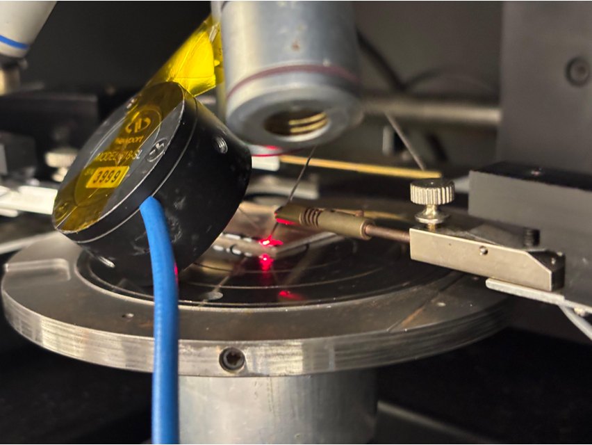 A probe contacting the laser die on the probe station, with red light visible and a photodetector positioned beside it