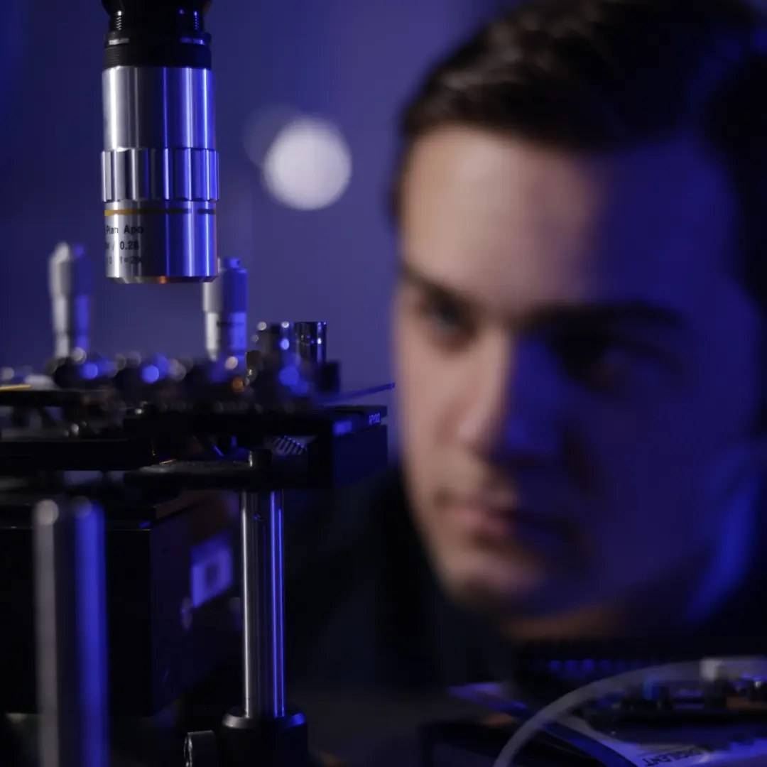 Microscope objective above the chip's alignment stage, with Tyler out of focus behind it