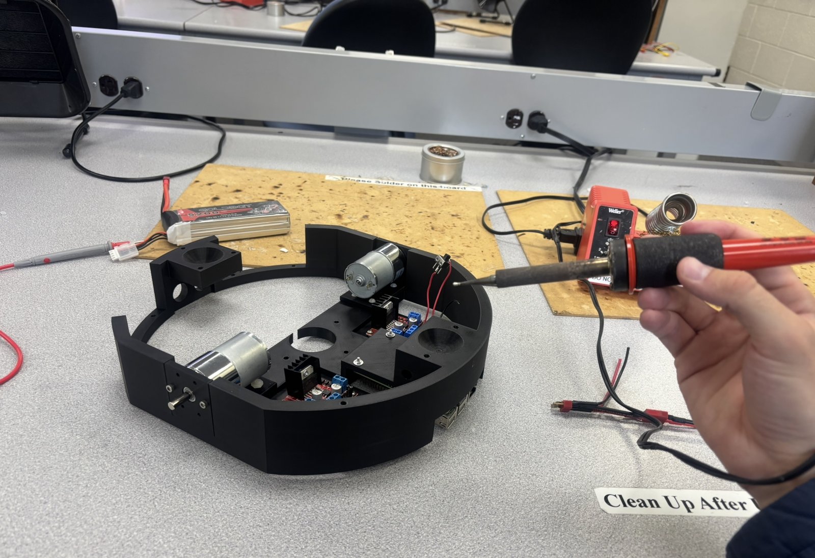 Soldering iron held over the partly assembled robot on a lab bench, with its motors, driver boards and a LiPo battery nearby